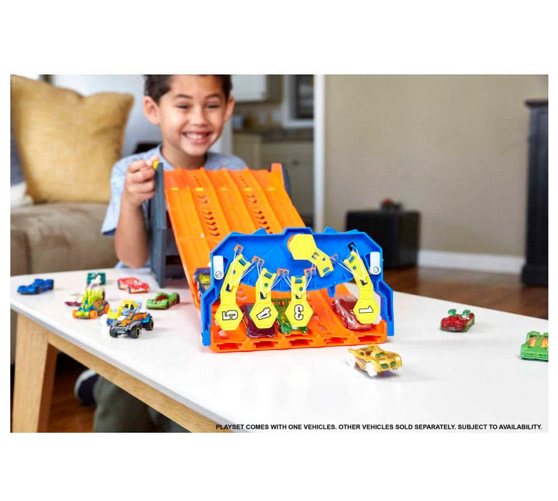 Excited boy in gray tshirt and end of orange plastic track, watches as hot wheels cars cross finish line. Finish line is the blue plastic end of track, with yellow number for each of the 5 tracks (1-5)--car flips up the number as it crosses the line. All on white table, scattered with Hot Wheels cars, 